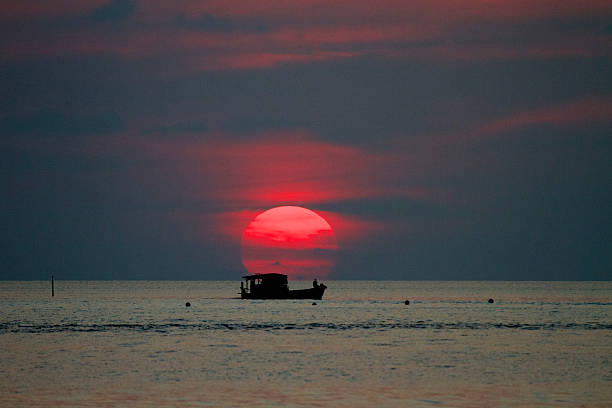 A big bright red sun sets over the ocean with a boat perfectly position in the foreground.