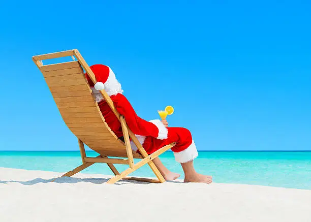 Christmas Santa Claus resting on wooden deckchair with fresh orange juice cocktail at ocean sandy tropical beach - New Year travel destinations  in hot countries concept