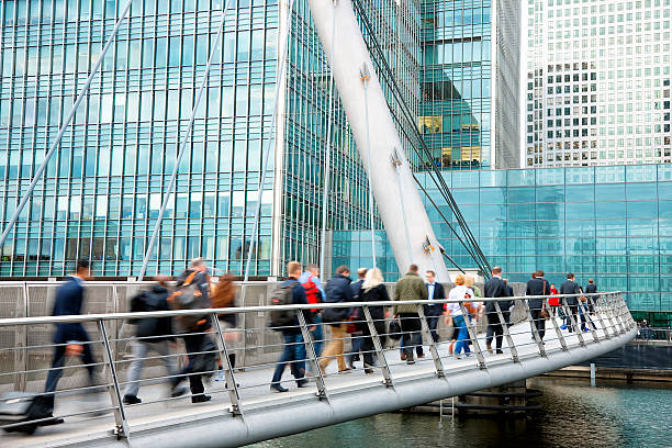 City Commuters Walking Across Bridge in Financial District, London, UK Crowd of city commuters, business people, office workers walking across suspension pedestrian bridge in London's financial district Canary Wharf. Blue, green and glass walls of financial buildings are seen in the background, Docklands, Isle of Dogs, London, England. canary wharf photos stock pictures, royalty-free photos & images