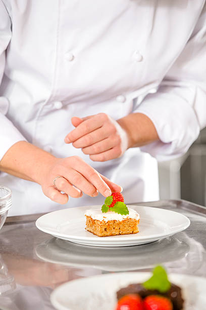 Close-up of a professional female chef who decorates dessert cake with strawberry and lemon leaf. Large industry kitchen. 