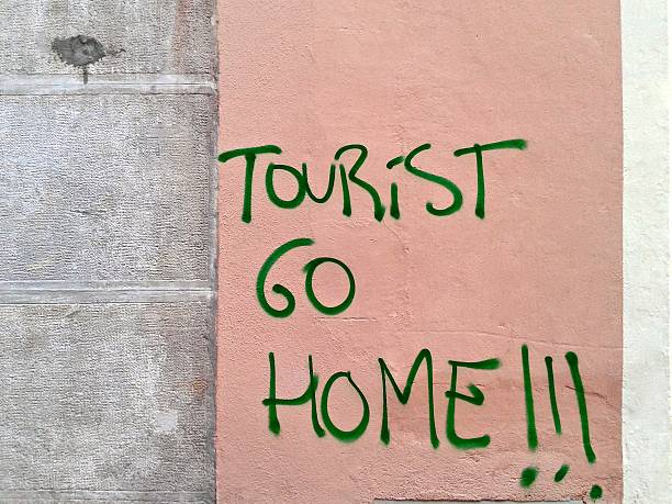Green spray-painted graffiti saying "Tourist Go Home!!!" on a pink wall beside gray stone tiles.