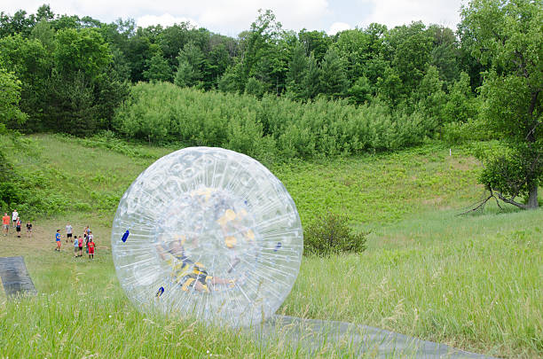 Zorb Rolling Down a Hill Lake Ann, Michigan, USA - July 1, 2014: a Zorb is a large sphere that 2 people ride in as it rolls down a hill. 2 young boy campers at Lake Ann Christian Camp, are strapped in and rolling down a hill in the Zorb as the rest of their group waits at the bottom of the hill to bring the Zorb back up for someone else to ride. zorbing stock pictures, royalty-free photos & images