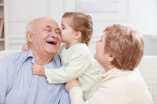 Pretty old men and women are spending time with their granddaughter. The girl is kissing her grandfather with joy. They are sitting on sofa and laughing