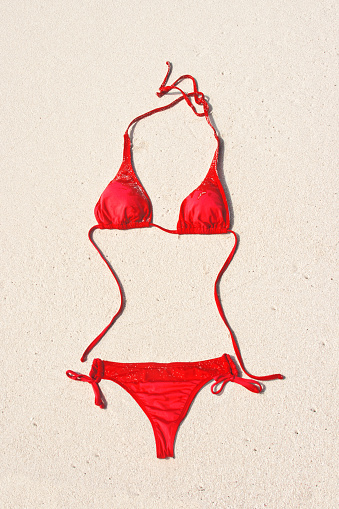 Red bikini swimwear lying on white sandy beach along Caribbean Sea at Cozumel, Mayan Riviera, Mexico. No people are around to claim the empty swimsuit, its strings shaped to form curves of a woman's figure. Playful image for idyllic tropical climate vacation at favorite travel destination.