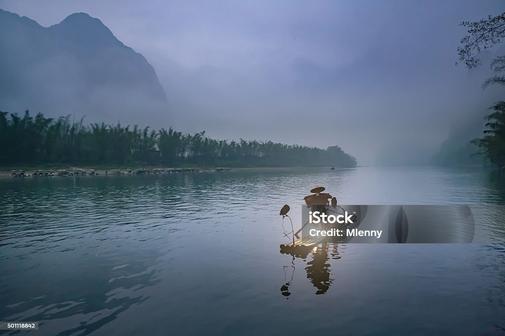 Li River Chinese Traditional Fisherman In Early Morning Fog Chinese senior fisherman in traditional clothes floating on his wooden raft with two gray herons on the Li River. Early morning fog at sunrise, fisherman is illuminated by his petroleum lamp. Tranquil, moody scene with real chinese fisherman in the early morning mist.  Xing Ping, Yangshuo County, Guangxi, Guilin, China. China - East Asia Stock Photo