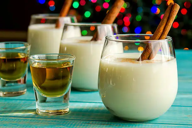 Row of small glasses filled with homemade egg nog with cinnamon sticks alongside shots of dark rum on blue wooden table with Christmas tree lights in background