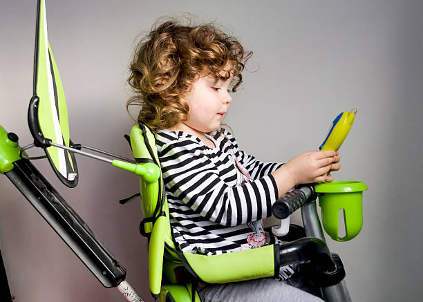 Young girl texting on bicycle 2 year old girl with curly brown hair, wearing a black and white striped top, texting or making a call with a mobile phone indoors on her green bicycle. toy phone stock pictures, royalty-free photos & images