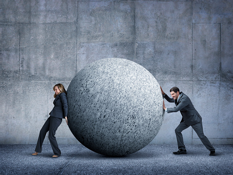 A businessman and a businesswoman working against each other while trying to move a large sphere as they both push against each other.