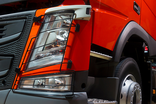 Headlight of oversize truck with stars, orange bodywork, bumper, hood, fender, door and wheel with silver disk of commercial cargo vehicle 