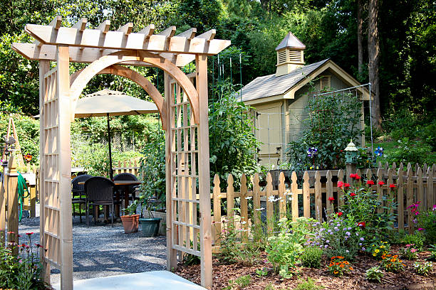 Garden Entrance and Potting Shed cedar wood arched trellis entry to a garden enclosed by a wooden picket fence with table and chairs and umbrella inside, potting shed off to the right with cupola trellis photos stock pictures, royalty-free photos & images