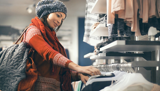 Closeup of smiling blond woman choosing clothes at department store in local supermarket. She's holding a beige blouse and looking at it. The woman is wearing gray cap, red sweater and scarf. Side view.
