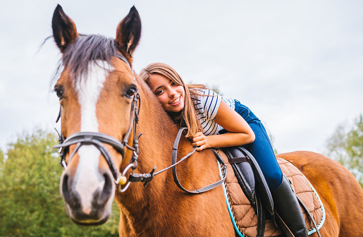 Happy young teenage girl leaning on the neck of her horse with a bright smile during an outdoor horseback ride in summer.