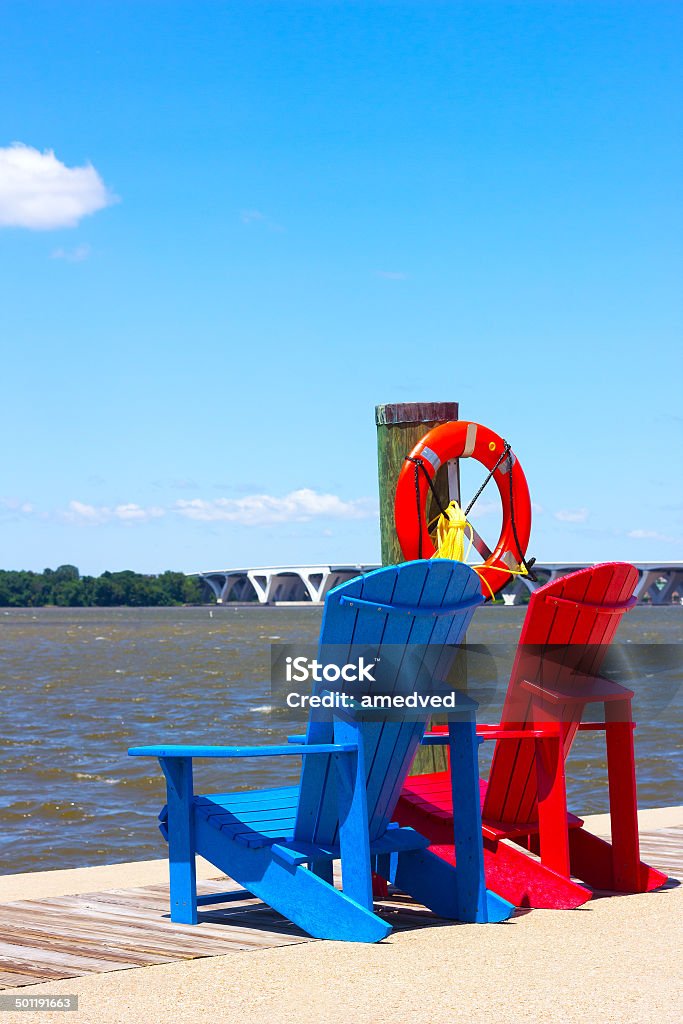 National Harbor pier with colorful chairs. Two wooden chairs and lifebuoy on the pier overlooking Woodrow Wilson bridge. Blue Stock Photo