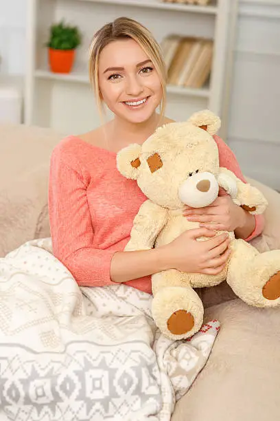 Cozy at home. Young pleasant woman is smiling while sitting on the sofa and hugging a teddy bear. 