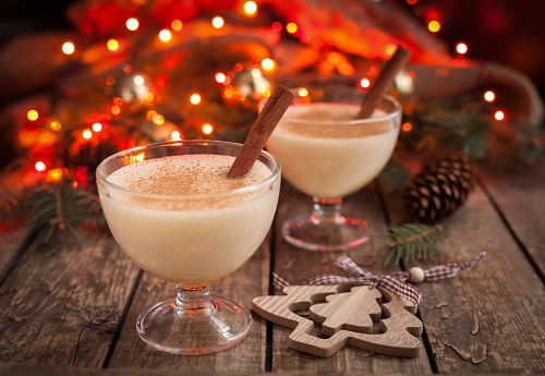 Eggnog traditional christmas egg, vanilla rum alcohol drink liqueur preparation recipe in two glass cups with cinnamon sticks on wooden vintage table. Red bokeh background. shallow depth of field.