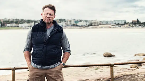 Color portrait of a mature, rugged, tough Australian man with Bondi Beach of Sydney in the background. 16x9 letterbox format with copy space on right.