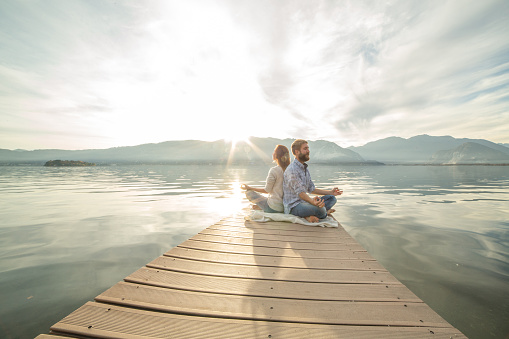 Young couple sits on a jetty above the lake, exercises yoga. Beautiful Autumn day in Italy.