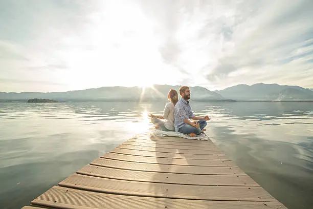 Photo of Young couple sits on jetty above lake, exercises yoga