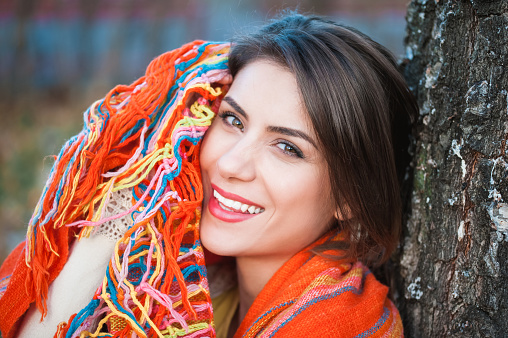 Portrait of young laughing  woman relaxing in beautiful evening in autumn park, next to a tree, holding a colorful scarf.