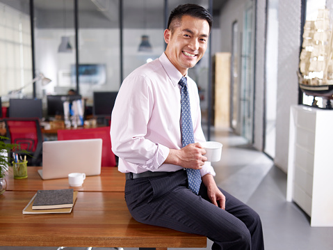 an asian businessman holding cup of coffee sitting on desk in office, smiling and cheerful.