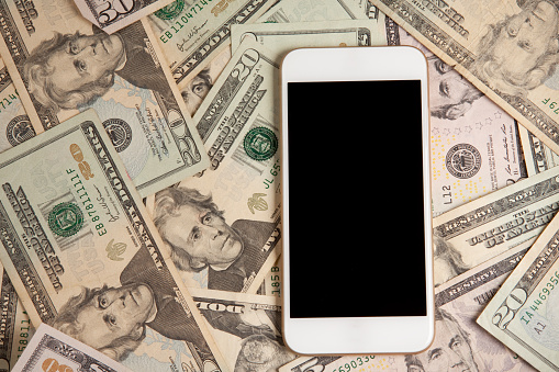An overhead view of a smart phone laying on top of a pile of US dollars.