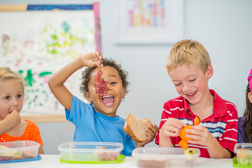 A multi-ethnic group of elementary age children are sitting at their desks and are eating their healthy lunches.