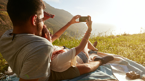 Rear view of young couple on picnic taking a self portrait with their smart phone.