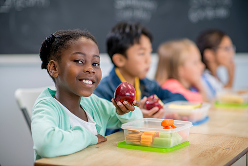 A multi-ethnic group of elementary age children are sitting at their desks and are eating their healthy lunches.