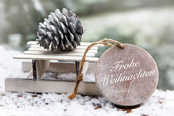 miniature wooden sleigh with pine cone and wooden tag with german 'Frohe Weihnachten' (Merry Christmas) in the snow