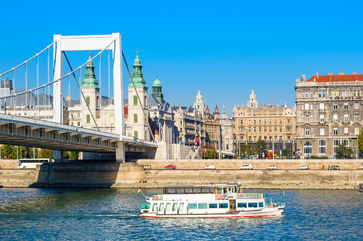 Photo of a tourboat passing by the Elisabeth Bridge on the Danube river with ornate buildings in the background in downtown Budapest Hungary on a blue sky day.