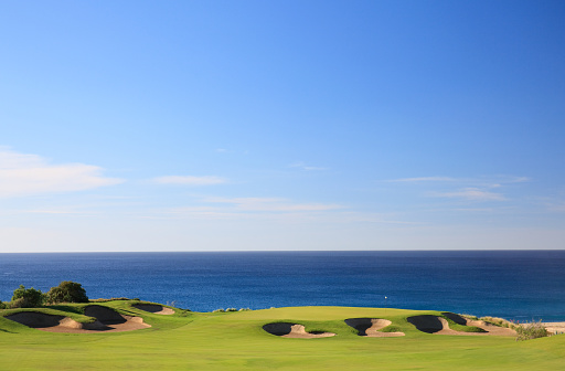 A beautiful image of a golf course in a tropical desert destination of Los Cabos, Mexico. Nobody is in the image. Image shows the beautiful topography of this stunning golf destination. The best in Mexico, perhaps the entire tropics! The flag is fluttering in the breeze and a beautiful residential area beside the Pacific Ocean. Classic golf course scenic with bunkering and excellent turf conditions and beautiful ocean backdrop behind the green.