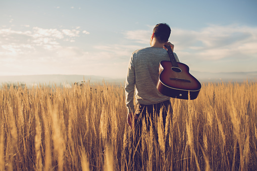 Rear view of a male guitarist standing in the middle of a prairie, enjoying a beautiful sunny day in the nature.