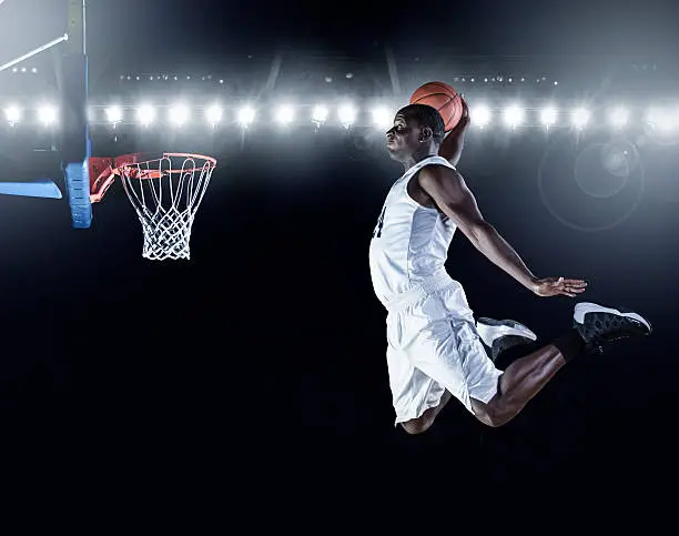 The side view of an african american basketball player scoring an amazing slam dunk in a professional basketball game. This is a composite image and not an actual basketball arena
