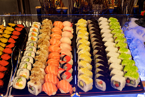 Close up macro color image depicting a delicious arrangement of fresh traditional Japanese sushi rolls topped with smoked salmon, prawn and avocado. There is a dish filled with soy sauce and also wooden chopsticks.