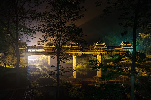 Dramatic backlit illuminated Wind and Rain Bridge in Chengyang at Night. The wind and rain bridge is a covered bridge in the traditional architecture of the Dong Minority Region and serves as link and meeting point for two villages.Light shining through the bridge column ofver the river. Silhouet trees in the foreground. Chengyang, Sanjiang County, Liuzhou, Guangxi, China