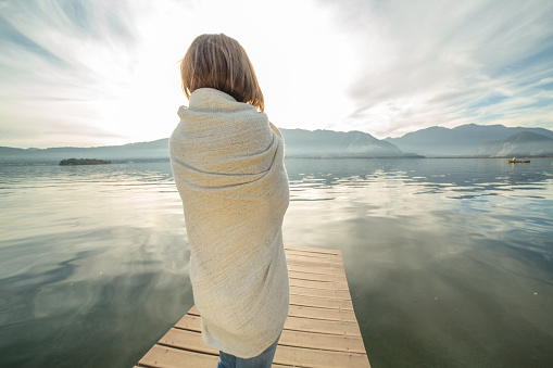 Young woman relaxes on lake pier, wrapped in blanket. Beautiful Autumn day in Italy.