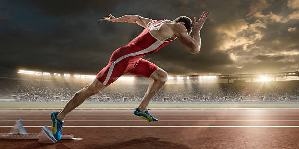 Male Sprinter Sprint Starts From Blocks in Athletics Stadium A close up image of a professional powerful male sprinter in mid action sprinting out of starting blocks at high speed, on a running track. The action takes place in a generic outdoor floodlit athletics stadium, full of spectators, under a dramatic evening sky. The athlete wears a bodysuit and running spikes.  track and field athlete stock pictures, royalty-free photos & images