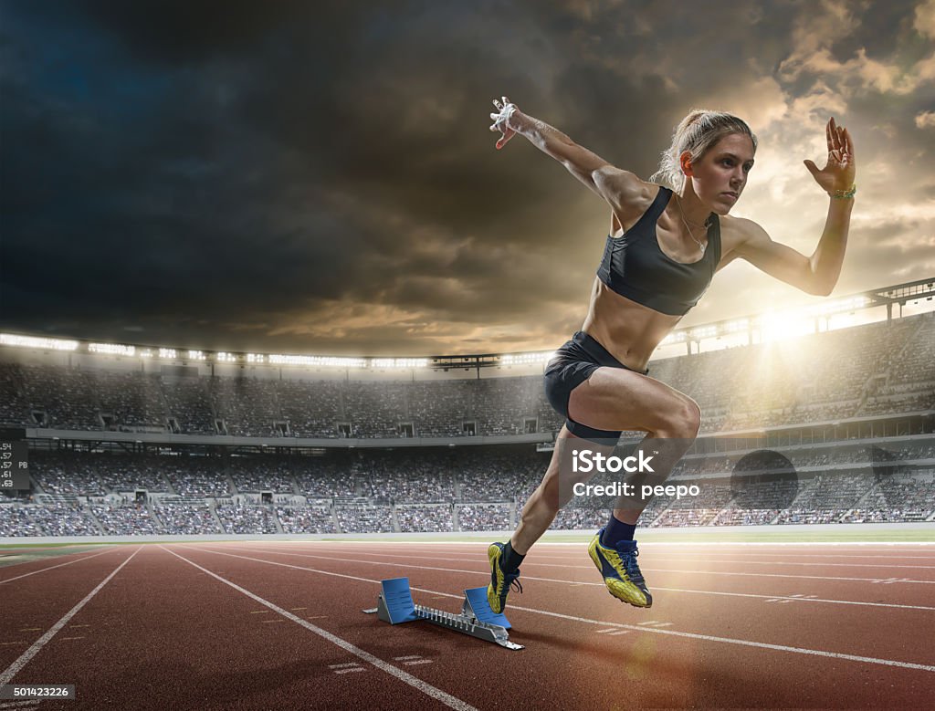 Woman Sprinter in Mid Action Bursting From Blocks During Race A mid action image of a woman sprinter during a sprint start from blocks on an outdoor athletics track. The athlete is running a generic outdoor floodlit athletics stadium full of spectators under a dark sky at sunset. The sprinter wears generic black sports top, shorts and running spikes.  Running Stock Photo