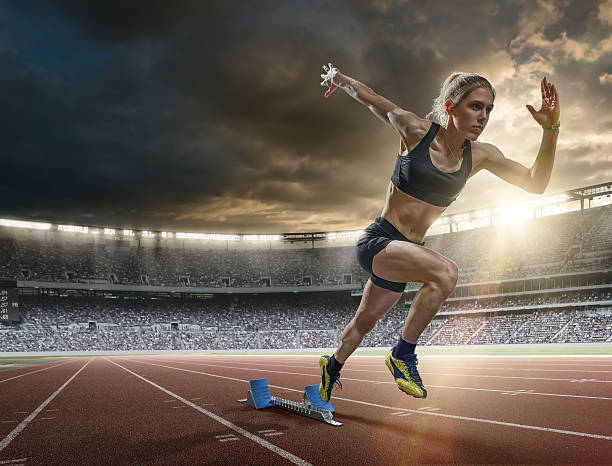 Woman Sprinter in Mid Action Bursting From Blocks During Race A mid action image of a woman sprinter during a sprint start from blocks on an outdoor athletics track. The athlete is running a generic outdoor floodlit athletics stadium full of spectators under a dark sky at sunset. The sprinter wears generic black sports top, shorts and running spikes.  womens track stock pictures, royalty-free photos & images