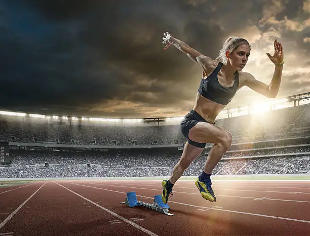 A mid action image of a woman sprinter during a sprint start from blocks on an outdoor athletics track. The athlete is running a generic outdoor floodlit athletics stadium full of spectators under a dark sky at sunset. The sprinter wears generic black sports top, shorts and running spikes. 