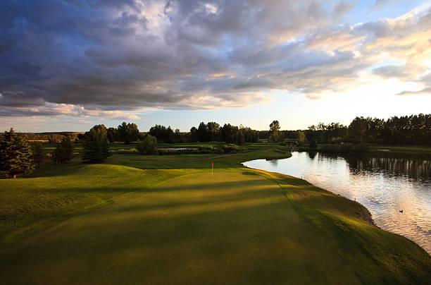 A beautiful golf course in a rural setting. Nobody is in the image. Beautiful golf course architecture with excellent turf conditions. Water hazard and beautiful mounding and shaping on a golf course. Pond is in the foreground. Green is backlit. Wide view of scenic golf course taken in mid summer. Image taken with aerial pole for high vantage view.