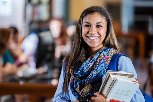Pretty mixed race high school or college female student is standing in modern public or school library holding a stack of textbooks. She is looking at the camera and smiling. She is wearing a printed scarf and backpack. Students are working at computers in the background.