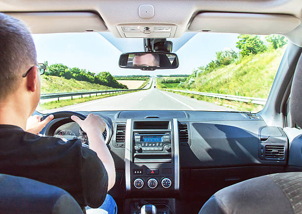 The man is driving his car with hands on the steering wheel. Long highway (highroad) is in the sunny summer day. Active caucasian male model. Travel (vacations), rental and adventure concept.