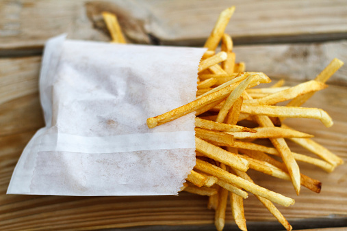 Shoestring French fries in a white bag on a yellow wooden picnic table.