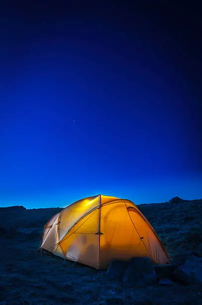 Stars starting to shine in a deep blue summer dusk sky over silhouetted mountain peaks and a bright yellow illuminated dome tent. ProPhoto RGB profile for maximum color fidelity and gamut.