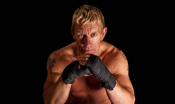 Serious Shirtless Old Fighter with Boxing Wraps A serious 60 year old male fighter with tattoos looking mean while wearing black boxing wraps with his arms propped up and hands together. The person is isolated on a black background. weight class stock pictures, royalty-free photos & images
