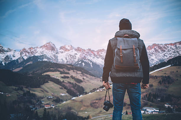 A man on vacation hiking in Austrian Alps. Wearing warm clothes, backpack and camera. He took a break to rest and enjoy in beautiful landscape. Winter day. Mountains are covered with snow. View from behind.