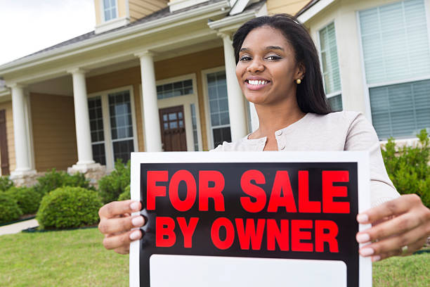 Young woman holding ""for sale by owner"" sign outside house Young woman holding "for sale by owner" sign outside house  house for sale by owner stock pictures, royalty-free photos & images