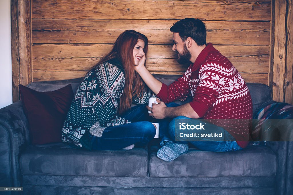 Hot drink and cozy sweater Couple sitting on sofa in wooden cabin. Wearing Christmas sweaters. Together enjoying in the warmth of their home for Christmas with cup of tea. Austrian Alps. Wooden wall in background. Adult Stock Photo