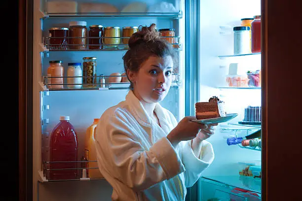 A young caucasian woman standing in front of the open refrigerator, eating unhealthy chocolate cake at late night in a domestic home kitchen. She is dressed in a casual bathrobe, looking at the camera with a surprised expression. A symbol of unhealthy eating habit and lifestyle. Photographed in vertical format.